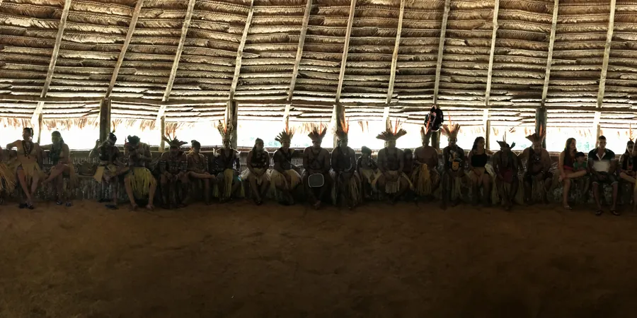 Integrantes de la comunidad, a contraluz, sentados en el banco de la maloca, la gran casa comunal de techo de hoja de palma