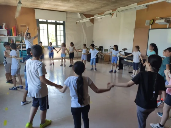 Alumnado de la Escola El Palau bailando en corro, cogidos de las manos, en la sala de la escuela; caras difuminadas