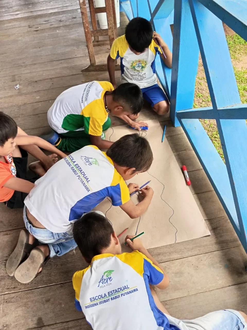 Niños puyanawa con el uniforme de la Escola Ixubãy Rabuī Puyanawa dibujan un río sobre una cartulina en el suelo