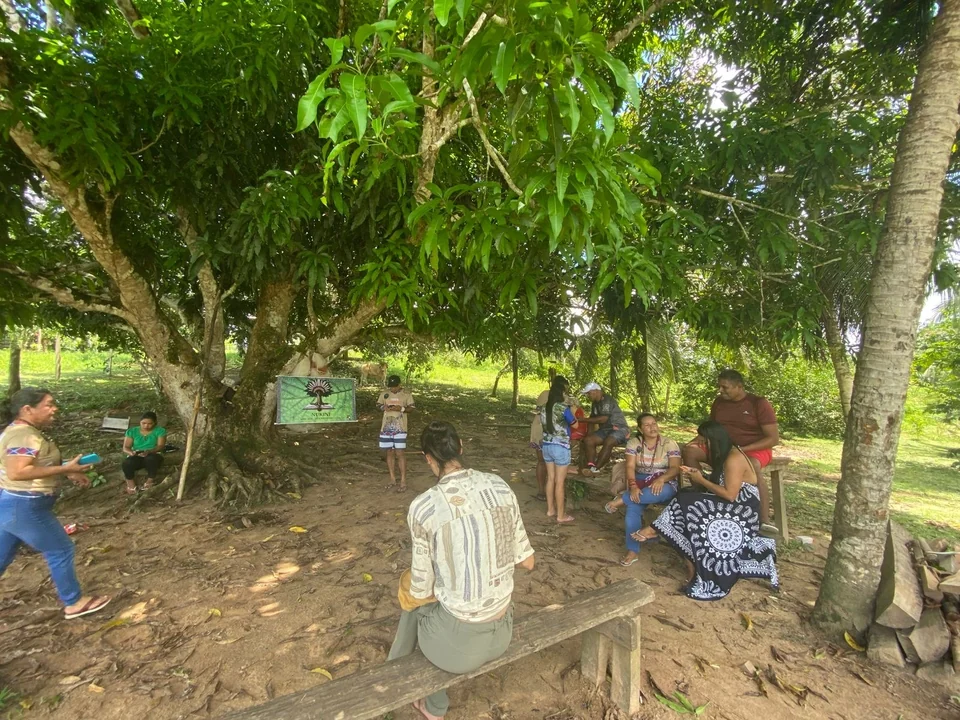 Un grupo de personas de la comunidad nukini sentadas a la sombra de un gran árbol, en la aldea