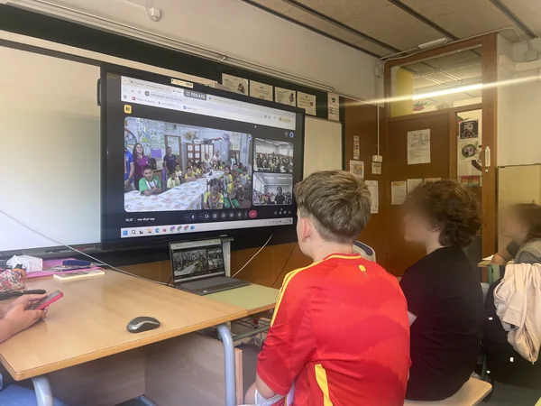 Alumnado de la Escola Sant Cristòfor, de espaldas, mira la pantalla del aula durante la videoconferencia con la escuela puyanawa