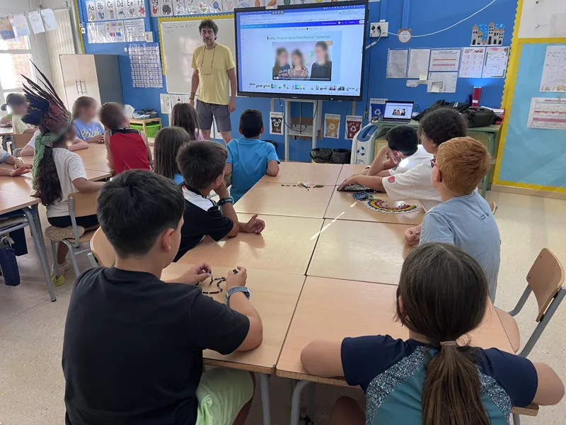 Taller de la Red en un aula de la Escola Sant Cristòfor de Begues: el alumnado, sentado alrededor de las mesas con artesanías amazónicas, escucha la presentación
